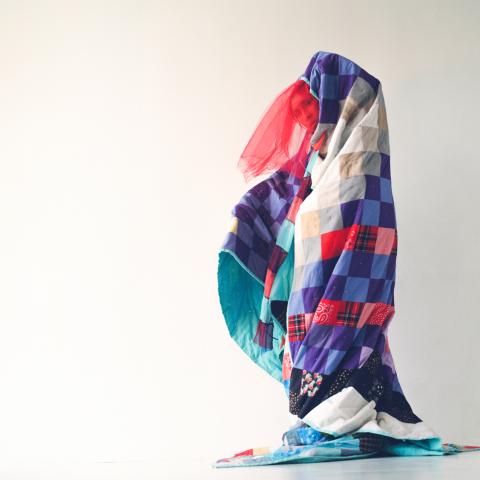 Under a multi-color quilt, a person peaks out. They pose in front of a white backdrop.