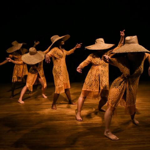 5 dancers standing in a diagonal row on a dim stage. They are wearing large woven hats and matching crochet dresses with arms out to their sides and looking in different directions.