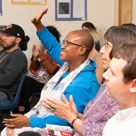 A woman in  bright blue clothing smiles as she raises her hand. She is seated in an audience in a conference room surrounded by other participants who are clapping.