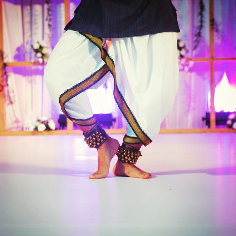A white floor with bare feet and brass ankle bells. A pink glow behind the dancer.