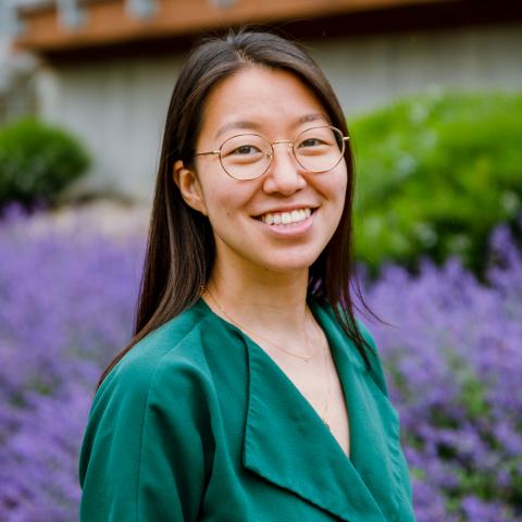 Jeena is an Asian woman. She wears a teal blouse and smiles in front of purple flowers. 