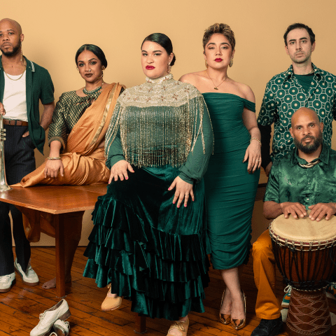 A group of six dressed in green pose in front of a mustard colored wall.