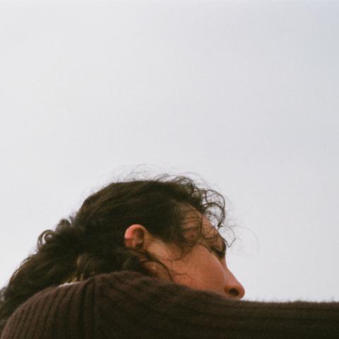 Maude's face is obscured by her shoulder as she turns away. She is Indigenous identifying and has long brown hair. She wears a brown sweater.
