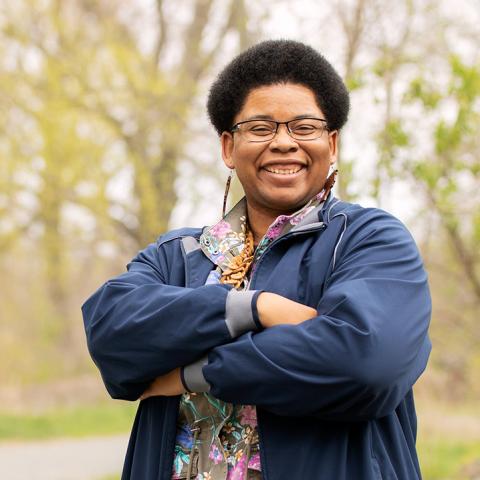 Outside next to a paved pathway, Catherine, who is a Black woman with a short afro, crosses her arms and smiles.