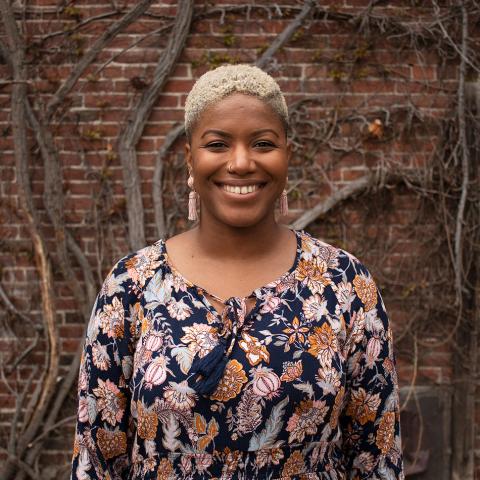 Christa is a woman of color. She has short bleach blonde hair and wears a floral dress. She poses in front of a brick building with vines.