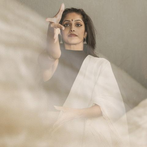 An Indian woman is behind a sheet fabric in the foreground. She holds her hand out and wears traditional garb.