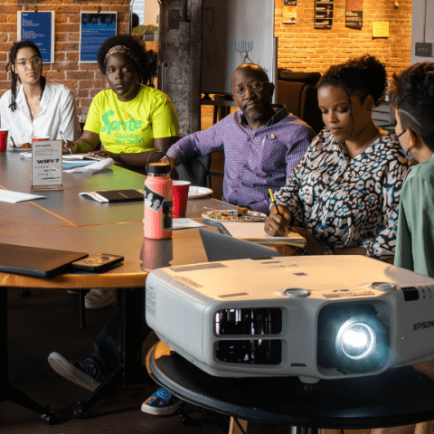 Eight folks sit around a table. A project is at the closer end of the table.