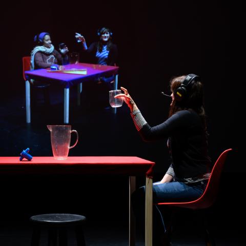 A person at a table holding a glass looking at a projection of  two people at another table holding glasses.