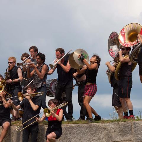 A dozen or more musicians playing brass instruments. A tuba player is leaping. They are outside on a granite-ledged park with a cloudy summer sky in the background.