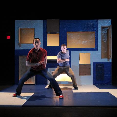  Two performers on a blue and white set. One performer slightly more in the foreground than the other.