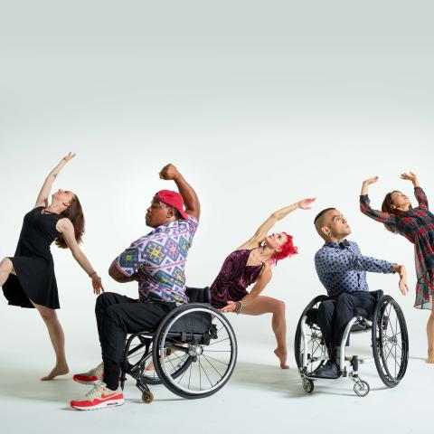 In front of a white backdrop, five dancers (two in wheelchairs) perform with their arms or legs out wide.