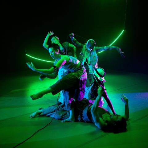 Four dangers, with their arms out and a couple of them in headpieces like helmets, perform with their arms wide and in front of a curved neon green light.