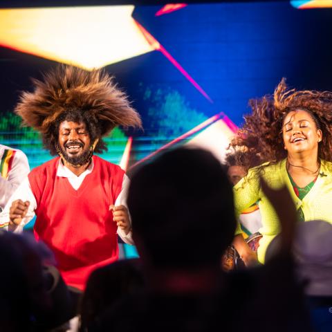 On a stage, and before a silhoutted audience, three folks, wearing bright colors, move their bodies and hit drums. A woman's hair is caught mid motion.