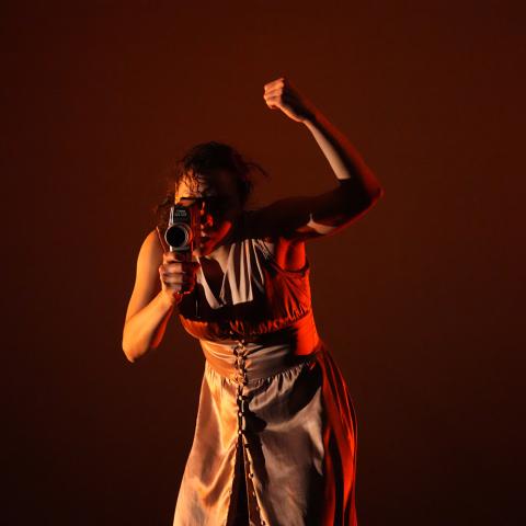 In dramatic red and yellow lighting, a light-skinned woman holds her fist up and looks through a super 8 camera lens.
