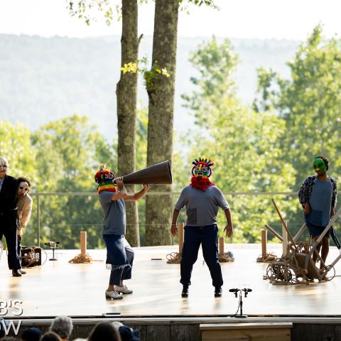 On a stage a group of masked performers observe a person in a bright mask.