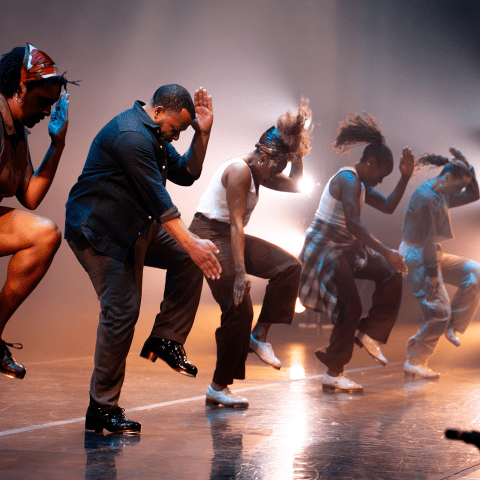Four performers on a row strike poses mid dance on a stage.