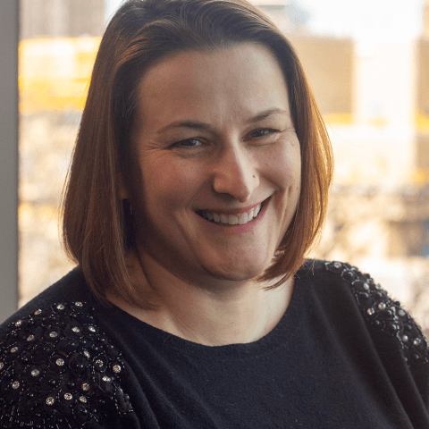 Adrienne has a brown bob. She is a white woman who wears a black sweater with rhinestones on the shoulders.