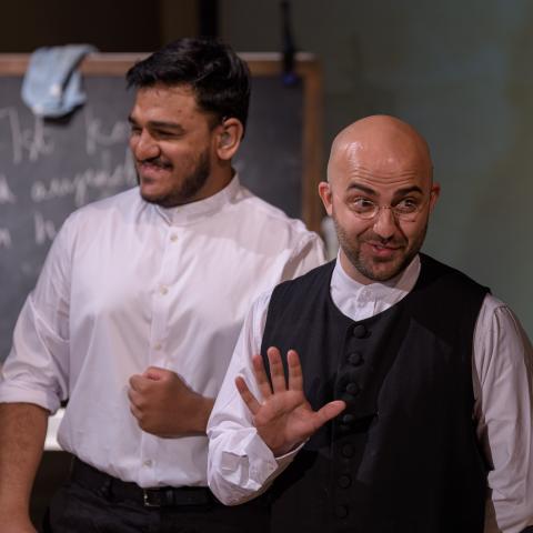 Brown men in white shirts stand in front of a chalk board in stage lighting.