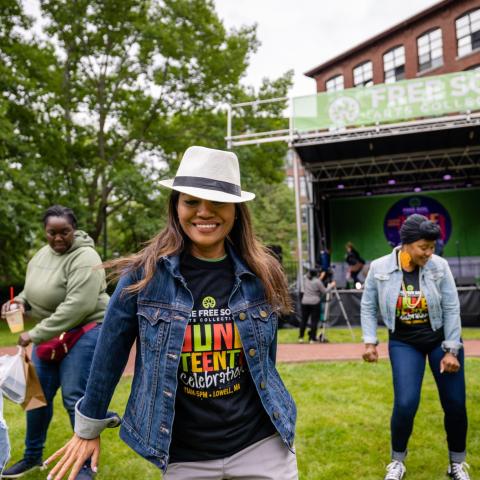 In a field, women of color, in event t-shirts, dance together. One of them, in the foreground, wears a fedora and smiles.