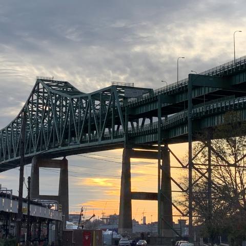 A view of the Tobin bridge from below with a cloudy sky with a bit of sun in the background