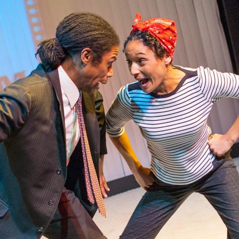 A black man in a suit and a black woman in a stripped t-shirt and red kerchief with arms akimbo leaning their heads toward each other