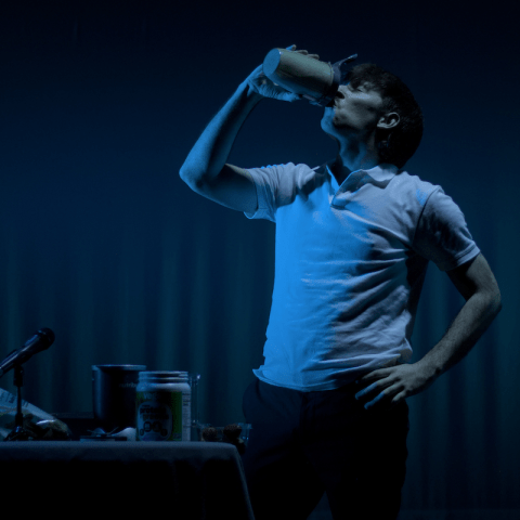 A person in a white polo shirt drinks from a mug in front of a dimly lit curtain.