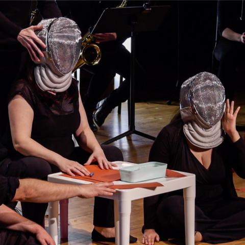 In stage lighting, three women in fencing or beekeeping masks sit around a table.