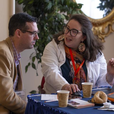 A woman in a doctor's costume gives a man in a suit a presentation on her laptop.