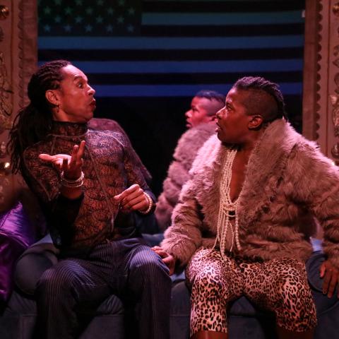 Two Black folk sit on a couch with stage lighting. They gesture to each other.