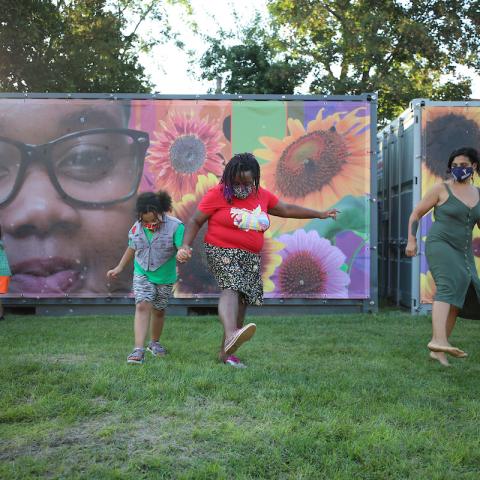 Two adults and two children dressed in brighly colored summer clothing dance on the grass in front of a mural. The sun shines through the trees in the background.