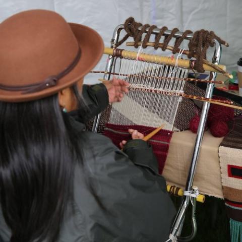 A woman, in a brimmed hat, weaves at a loom.