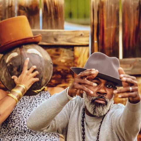 A Black woman holds a mask over her face, while wearing a top hat, and, in front of her, a Black man, in a fedora, holds his hands up.