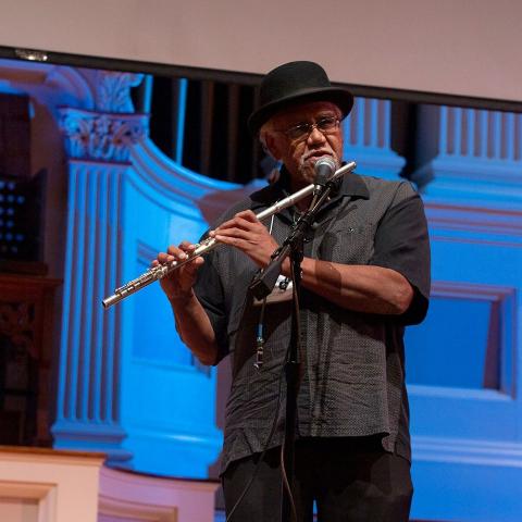On a stage, a man in a top hat plays the flute.