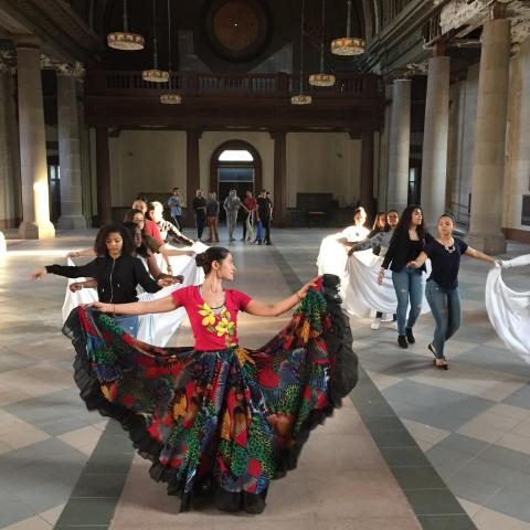 Woman rehearsing a dance in a church space. The woman in front wears a brighly colored long, wide skirt, and is holding up the edges up with each hand while looking back at others who seem to be doing the same.