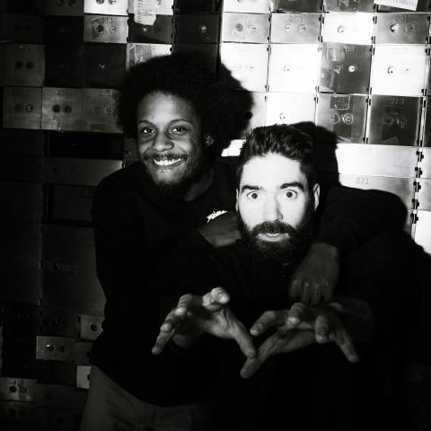 Black and white photo: a Black man and a white man pose against a metal wall in flash lighting.