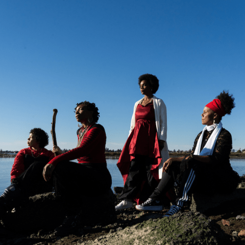 Four people are outside by a waterfront, three of them sit and one of them stands.
