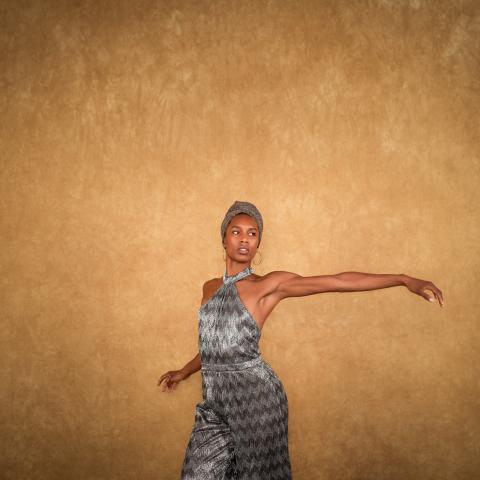 In front of a tan backdrop, a Black woman, in a grey headwrap and matching satin jumpsuit, holds her arm out to her left.