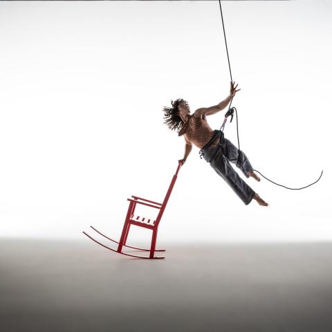 A person suspended in midair by a harness reaches upward with their left hand while touching a red rocking chair with their right.
