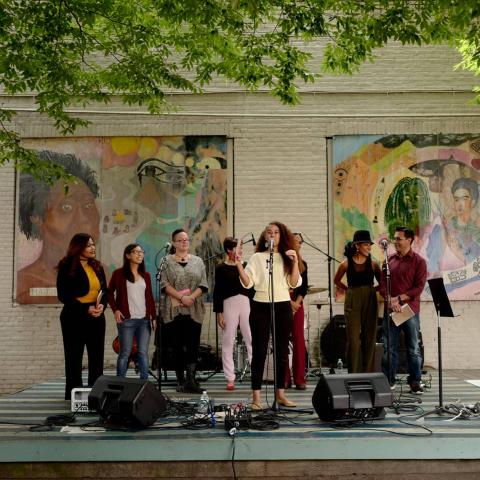 A woman, on stage, with other women presents her project, in front of a wall of murals of powerful women throughout history.