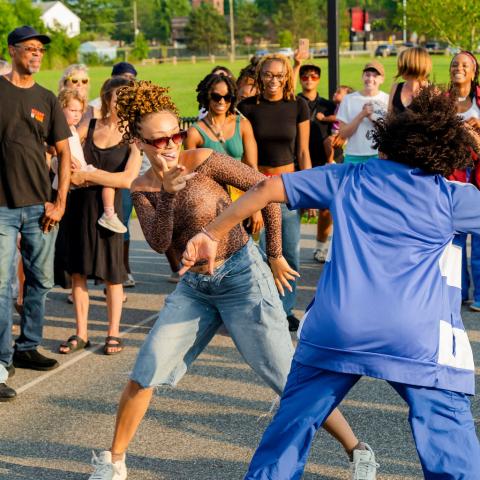 Outside, on a basketball court, two Black women dance among a circle of folks who watch.