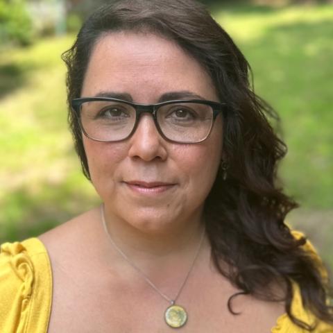 A woman with long dark hair, glasses, a gold pendent and a yellow blouse.