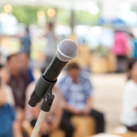 Outside, a standard black and silver microphone on a stand. Blurry folks congregate behind it.