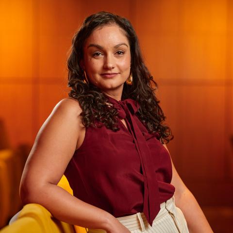 A brown woman with shoulder length, curly brown hair wears a sleeveless, maroon blouse and white pants. She is in an auditorium with yellow chairs and wood panelling. 