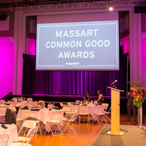 An empty gala hall with a projection of "MassArt Common Good Awards" in front of a stage.