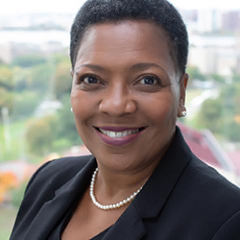 A head shot of awoman in a black suit jacket and pearls; the background appears to be a park.