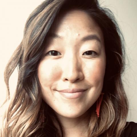 A woman of Asian descent with long wavy hair, wearing a black top and red earrings against a neutral background.