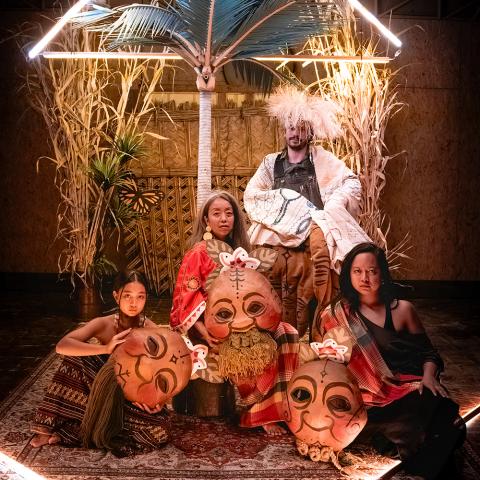 Four performers sit together under and behind neon lights. Three women hold large masks and a man sits behind them in a garment with wide and long sleeves.