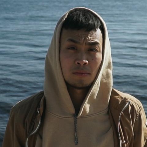 A man with short dark hair wearing a tan hoodie with a body of water in the background.