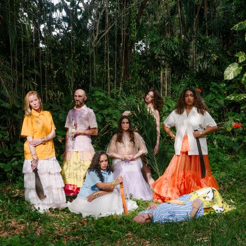 7 dancers wearing guayaberas and long skirts are arranged kneeling, laying, crouching, and sitting in a tropical outdoor setting. They look into the camera with varying expressions of intensity and devotion. Some carry machetes or other symbols of cuban American iconography.