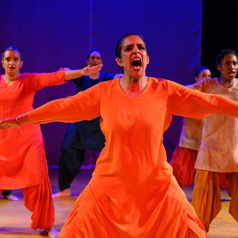 A brown woman in orange dress holds her arms out and screams. Four dancers are behind her.